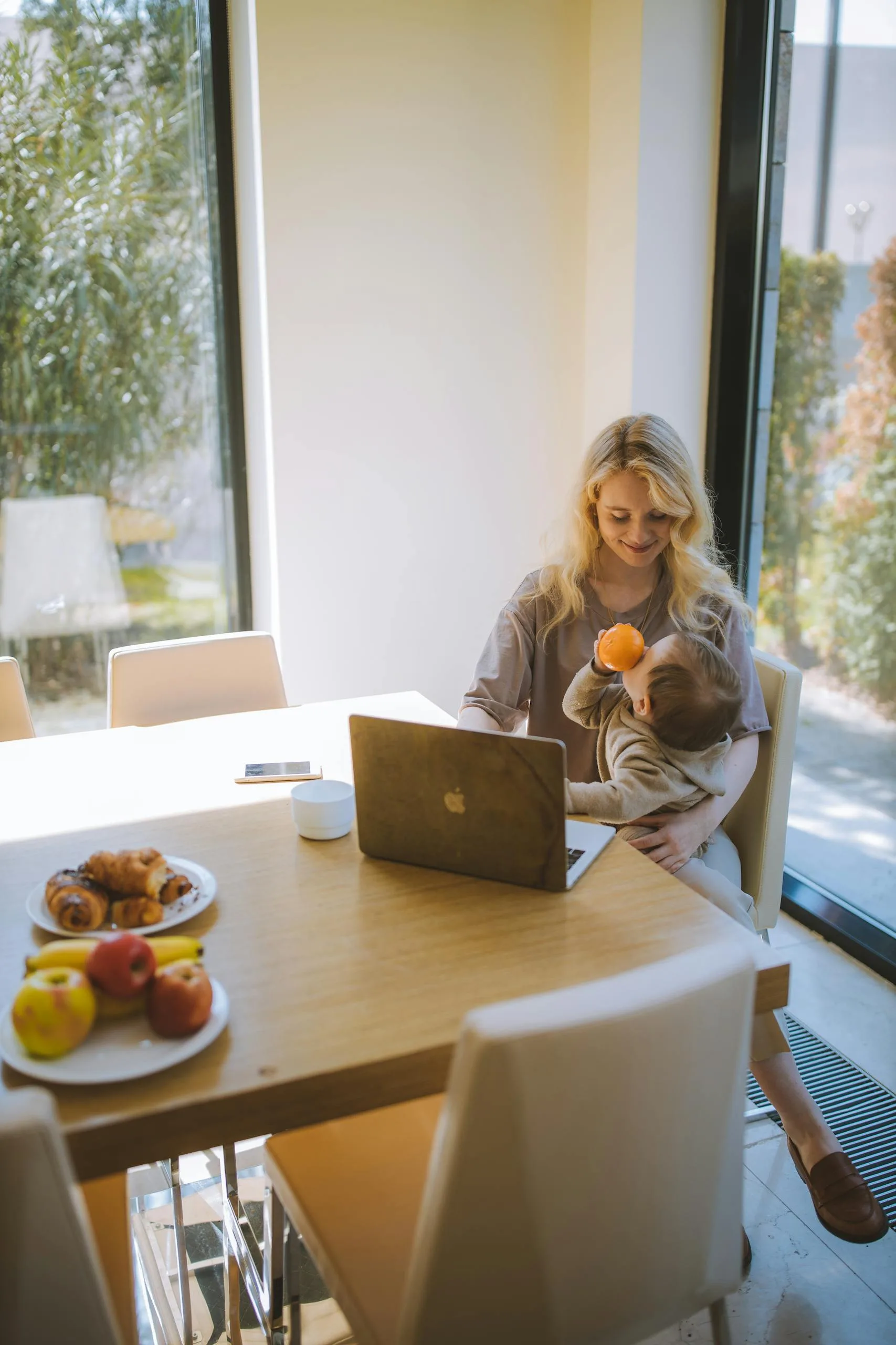 A blonde mother multitasks with her baby and laptop, enjoying breakfast in a bright home setting.